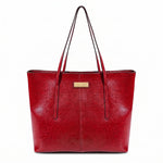 A red faux-leather tote bag with dual shoulder straps and a small gold-tone metal brand plaque on the front, shown against a plain white background.