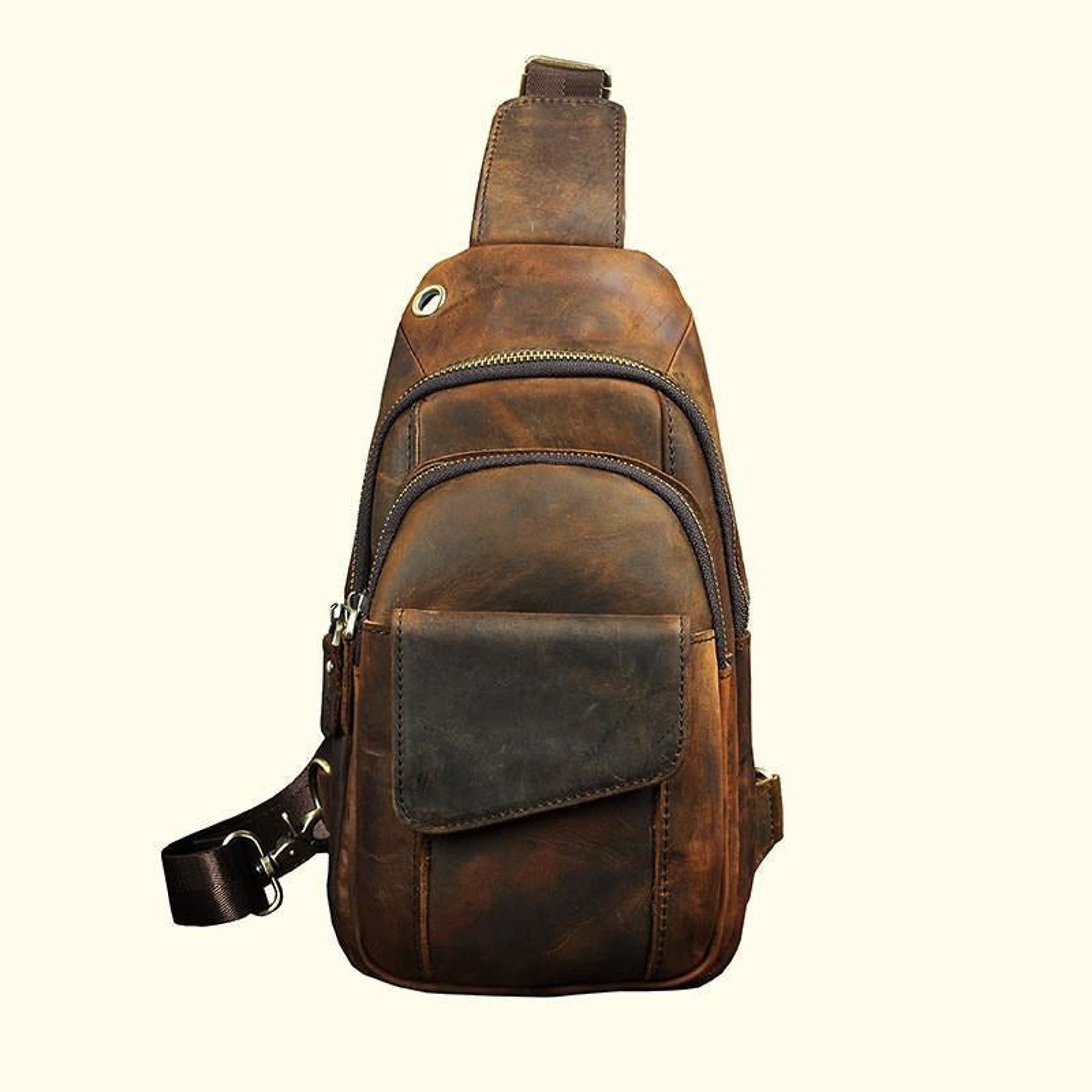 A brown distressed leather sling backpack with a front pocket and adjustable shoulder strap, shown against a plain cream background.
